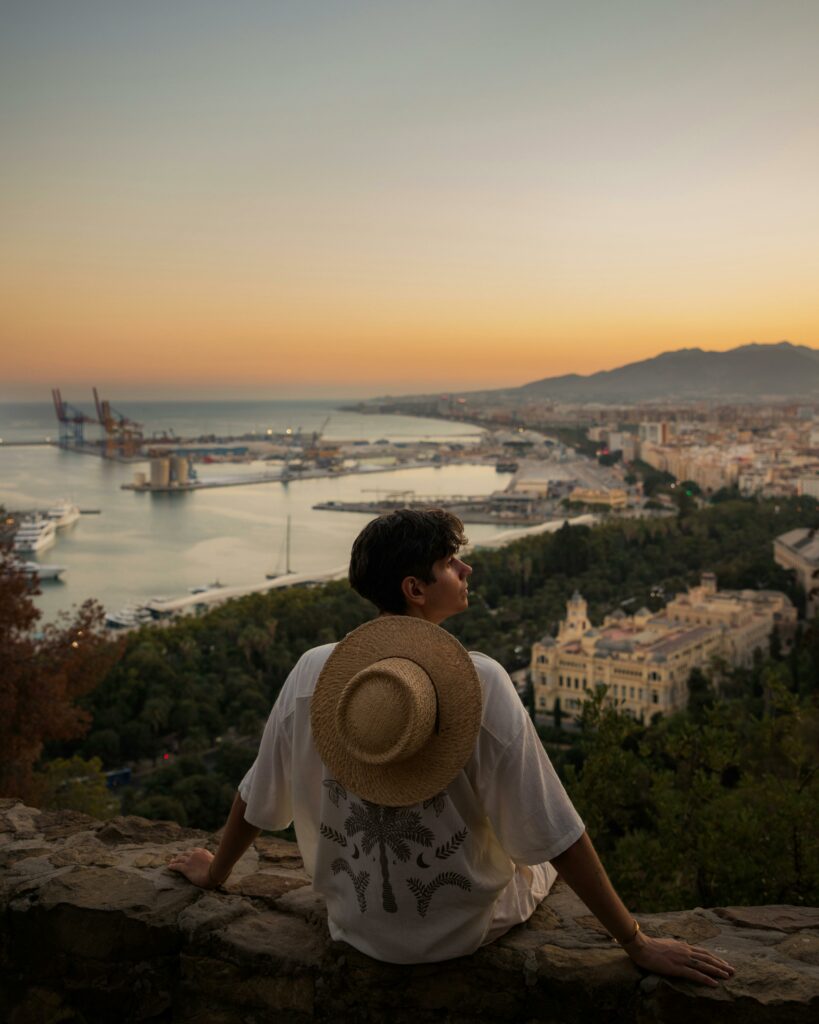 Man in hat overlooking Málaga cityscape and harbor at sunset.